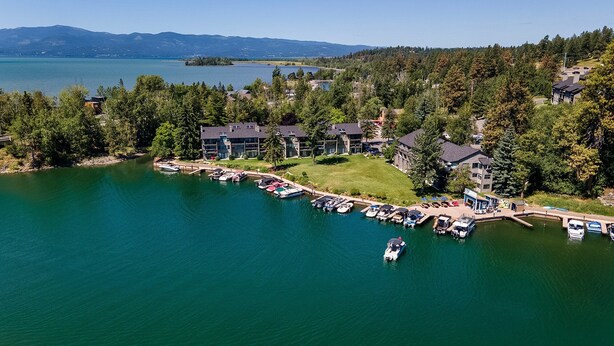 An aerial view of a lakeside property with multiple buildings, a dock with boats, and a clear view of the lake and mountains in the background. - Stunning views of the lake, private balcony & gorgeous fireplace - Spacious Marina Cay Getaway (Bigfork)
