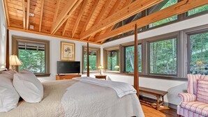 A bedroom with a large bed, wooden ceiling beams, multiple windows, a television, a bedside table, and a chair. - High Haven | Breathtaking Mountain Views! (Black Mountain)
