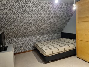 Een slaapkamer met behang met een patroon, een bed met een geruit sprei, een houten garderobe en een plafondlamp.