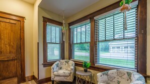 A room with wooden floors, a large window with blinds, a chair, a small table with a plant, and a hanging plant. - Remodeled Rocheport Sunflower House - A Bungalow in Historic Rocheport (Rocheport)