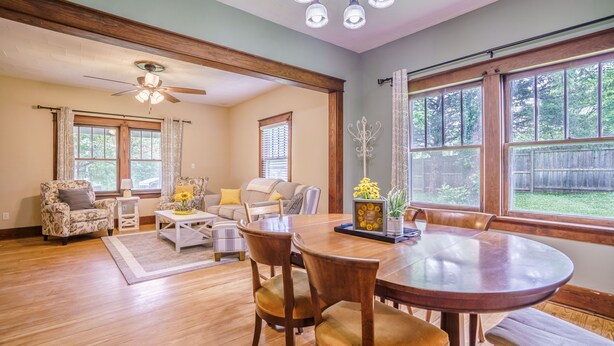 A spacious living room with a wooden floor, a ceiling fan, a dining table with chairs, and large windows offering a view of the backyard. - Remodeled Rocheport Sunflower House - A Bungalow in Historic Rocheport (Rocheport)
