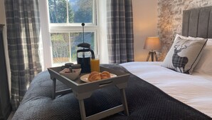 Una cama bien tendida con un cojín decorado con un diseño de ciervo, una lámpara de noche y una bandeja con cruasanes, una cafetera y un vaso de jugo de naranja. La cama ofrece vistas a árboles a través de una ventana.