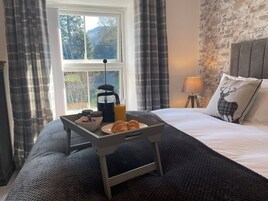 A neatly made bed with a pillow featuring a deer design, a bedside lamp, and a tray with croissants, a coffee pot, and a glass of orange juice on a bed with a view of trees through a window.