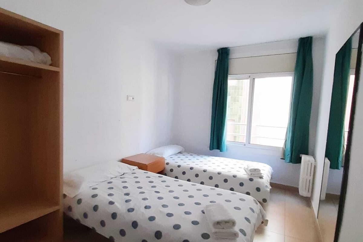 A small, clean room with two beds, a wooden wardrobe, a radiator, and a window with curtains.