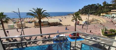 A balcony with a pool, beach view, palm trees, and a parking area.