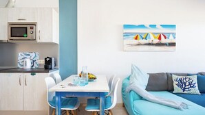 A modern kitchen with a microwave, coffee maker, and kettle. There is a dining table with chairs and a painting of a beach scene. A teal sofa with a decorative pillow is also visible. - Home2Book Bright Penthouse Médano, Terrace & Pool (El Medano)