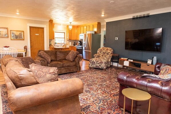 A living room with a sofa, armchair, and ottoman, a flat-screen TV, and a kitchen in the background. - River House in historic Rockton, IL (Rockton)