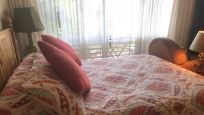 A bed with a patterned bedspread and two red pillows, a green floor lamp, and a view of a balcony through sheer curtains. - Lodging Near Public Transportation (Acton)