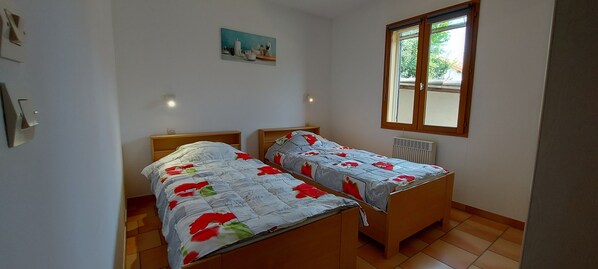 Een slaapkamer met twee houten bedden, een radiator, een raam met uitzicht op groen, en een klein schilderij aan de muur.