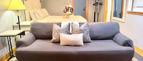 Una habitación de hotel moderna con un sofá gris, una mesa de centro de madera, una cama con cabecero blanco y un espejo en la pared.