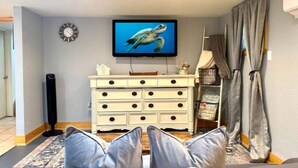 Een woonkamer met een witte dressoir, een televisie waarop een schildpad wordt getoond, een klok en een raam met gordijnen.