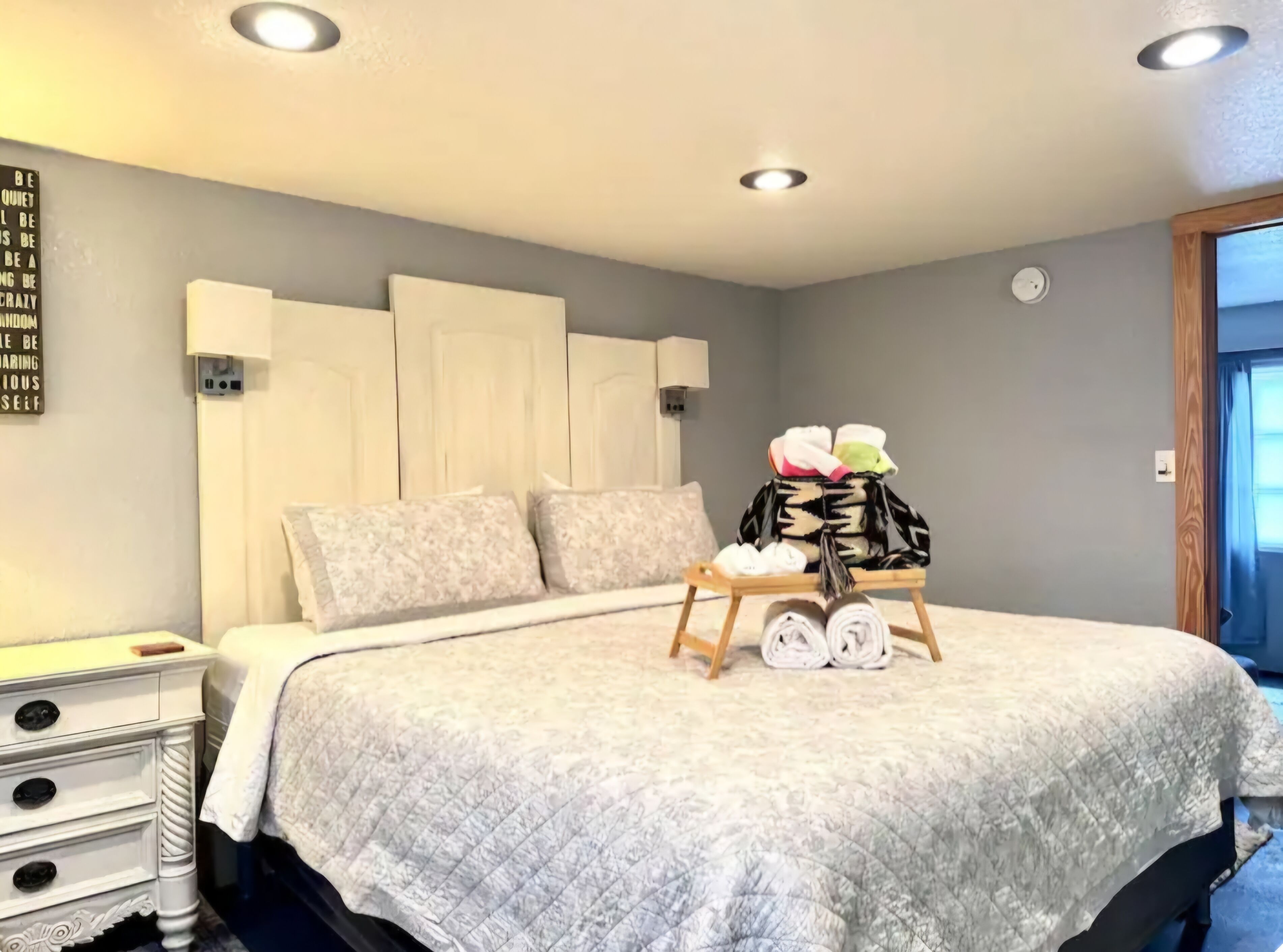 A neatly made bed with a quilted blanket, a bedside table with drawers, a wall clock, and a bag of folded clothes on a small wooden stand.