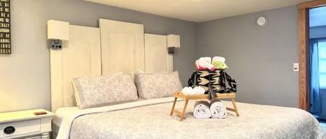 A neatly made bed with a quilted blanket, a bedside table with drawers, a wall clock, and a bag of folded clothes on a small wooden stand.