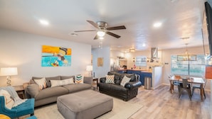 A spacious living room with a grey sofa, a blue armchair, and a black leather couch. There is a dining area with a table and chairs, and a kitchen in the background. - Trendy Pet-friendly House w/ Patio near Lake LBJ (Kingsland)