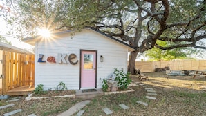A small white building with a pink door, labeled "Lake," surrounded by a wooden fence and a large tree. - Trendy Pet-friendly House w/ Patio near Lake LBJ (Kingsland)