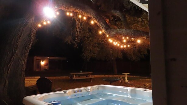 An outdoor hot tub area with a tree and string lights, a picnic table, and a small table with a bird feeder. - Trendy Pet-friendly House w/ Patio near Lake LBJ (Kingsland)