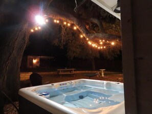 Un espace extérieur aménagé avec un bain à remous, un arbre et des guirlandes lumineuses, une table de pique-nique et une petite table sur laquelle est posée une mangeoire à oiseaux.