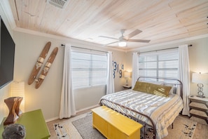 Une chambre à coucher comprenant un lit, un ventilateur de plafond, une fenêtre avec des stores, une table de chevet et une lampe.