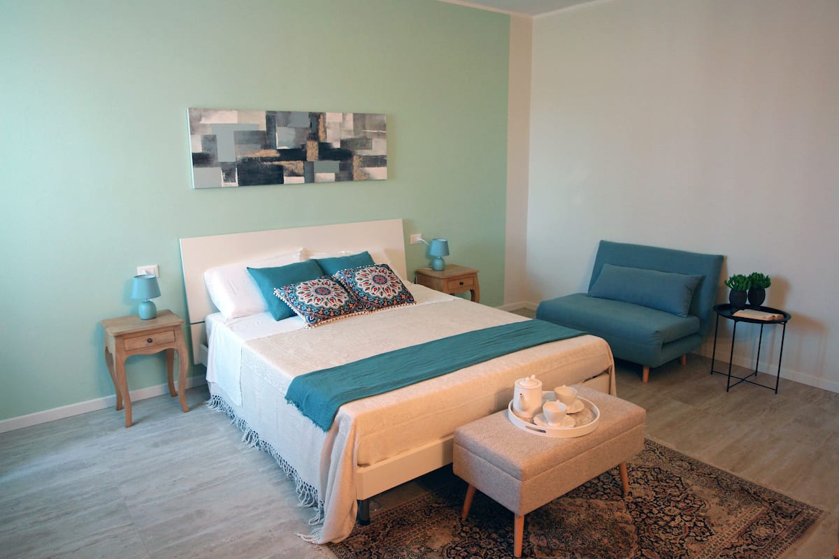 Une chambre à coucher comprenant un lit, des tables de chevet, une chaise, une petite table avec un plateau, et une œuvre d’art abstraite accrochée au mur.