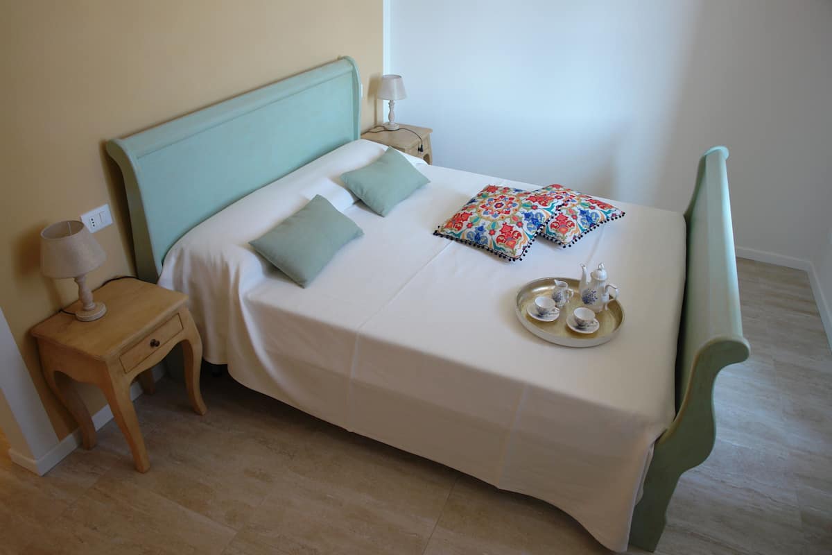 Un lit bien fait, recouvert d’une couvre-lit blanche et agrémenté de deux oreillers, une table de chevet avec une lampe, et un plateau avec de la vaisselle à thé posé sur le lit.