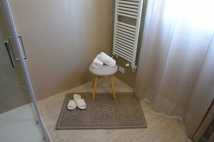 Un bagno con doccia, un asciugamano su uno sgabello e ciabatte per terra.
