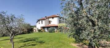 Una casa a due piani con tetto in tegole rosse, circondata da un prato curato e alberi di ulivo.