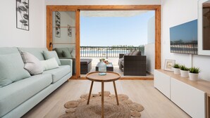 A modern living room with a light green sofa, a round wooden table, and a view of a balcony with outdoor furniture. - BEACHSIDE CARABELA APARTAMENTOS (Alcúdia)