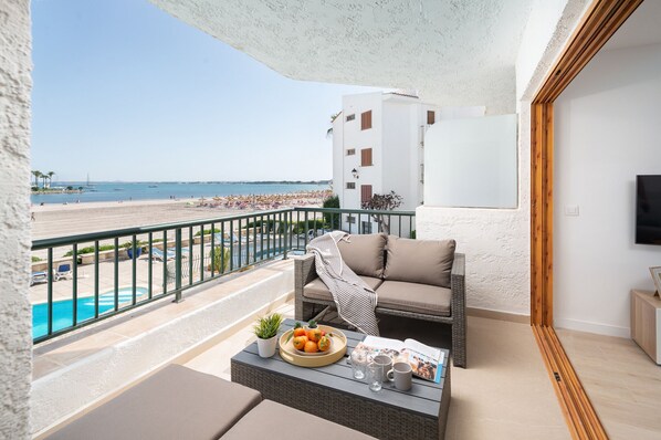 A balcony with a wicker sofa, a small table with fruits and drinks, a pool, and a beach view. - BEACHSIDE CARABELA APARTAMENTOS (Alcúdia)