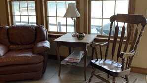 A living room with a leather sofa, a wooden rocking chair, a small table with a lamp, and a view of a lake through large windows with red curtains. - Railway camp on Moosehead Lake (greenville)
