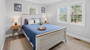 Een slaapkamer met een houten bed, twee nachtkastjes, een wandlamp en twee kunstwerken aan de muur, met uitzicht op bomen.