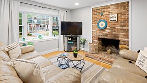 Een woonkamer met een leren bank, een flatscreen televisie, een metselwerk schoorsteen en een groot raam met gordijnen.