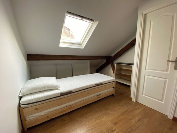Une petite pièce avec un plafond en pente, comprenant un lit simple, un puits de lumière et une porte.