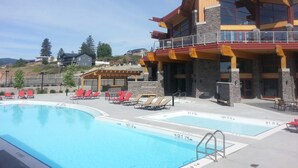 A pool area with red lounge chairs, a stone building with balconies, and a clear sky. - Okanagan four seasons paradise with shared pool and hot tub (Westbank)