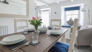 En matplats med ett träbord dukat för fyra personer, vita stolar, en vas med blommor och utsikt över ett vardagsrum med en soffa och ett fönster.