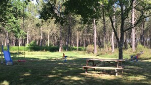 Un área de picnic con mesa y bancos, rodeada de árboles y un parque infantil con tobogán y columpios.
