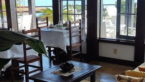 Amplia habitación con grandes ventanales que ofrecen vistas al mar. Hay un sofá blanco con cojines estampados, una mesa de centro de madera oscura y una pequeña mesa con mantel blanco y sillas.