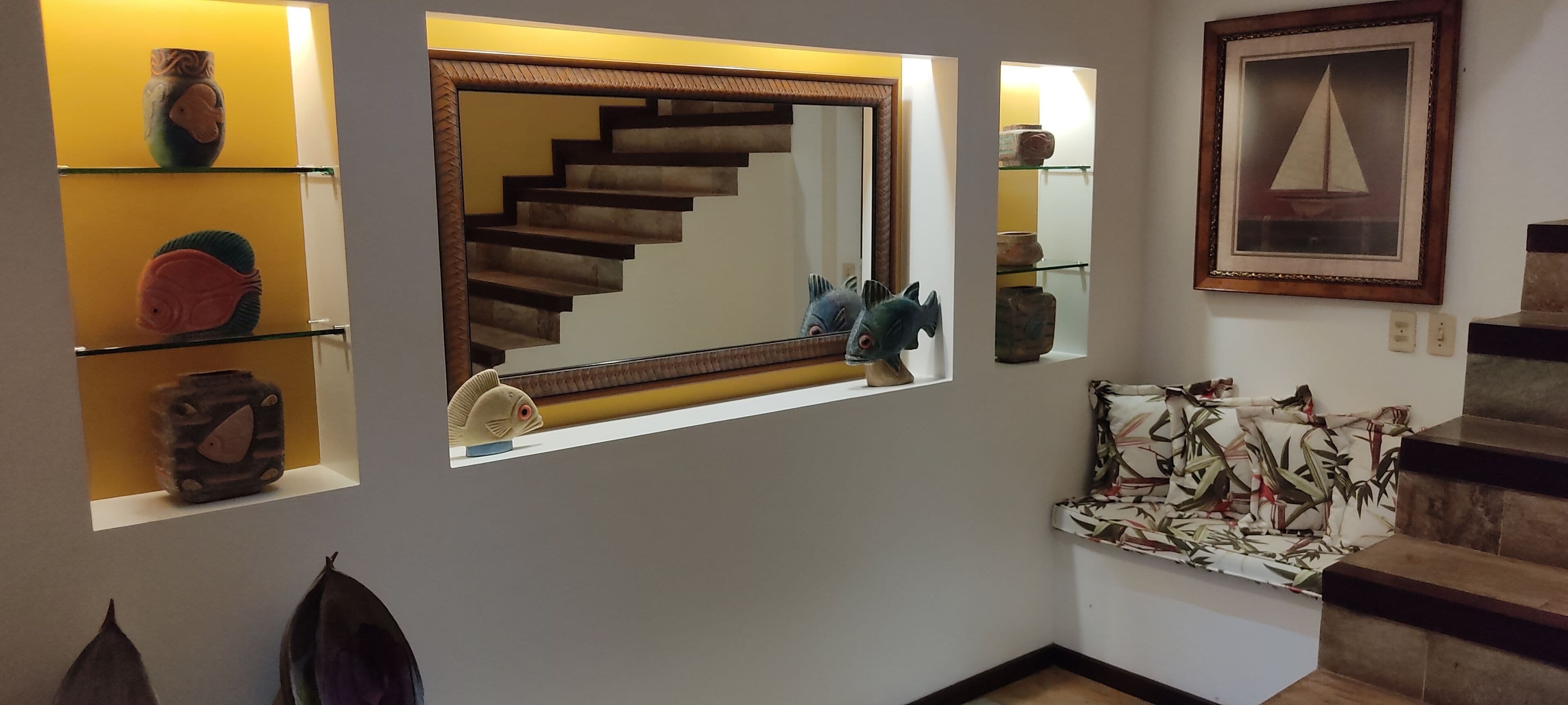 Una escalera con acabado de madera, un espejo que refleja una escultura de pez, y estantes con adornos.