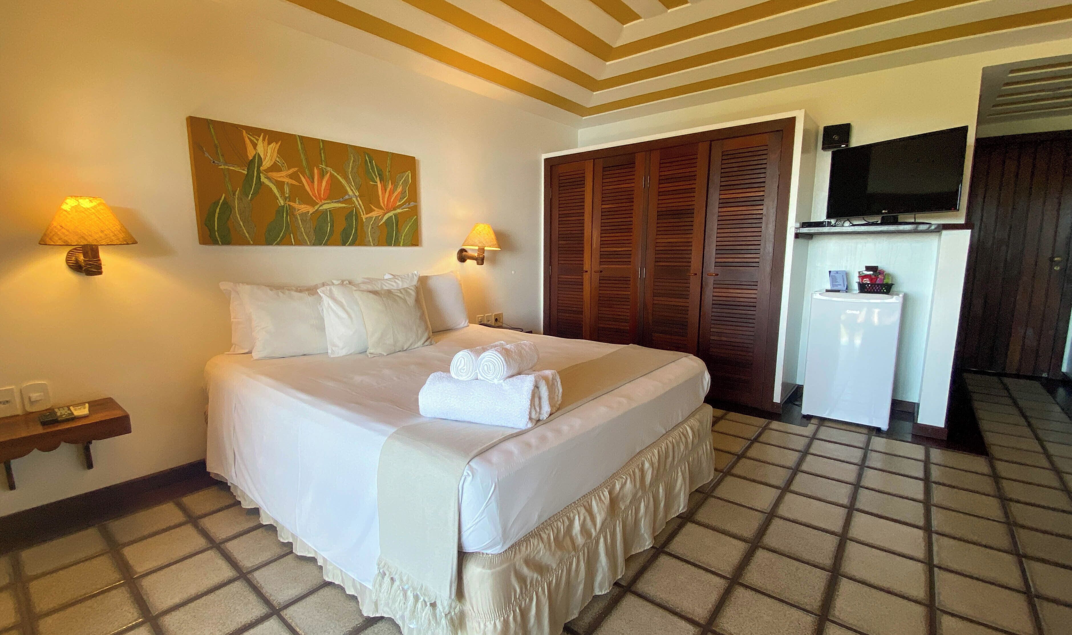 A neatly made bed with white linens and two rolled towels, a wall-mounted TV, and a small refrigerator in a hotel room.