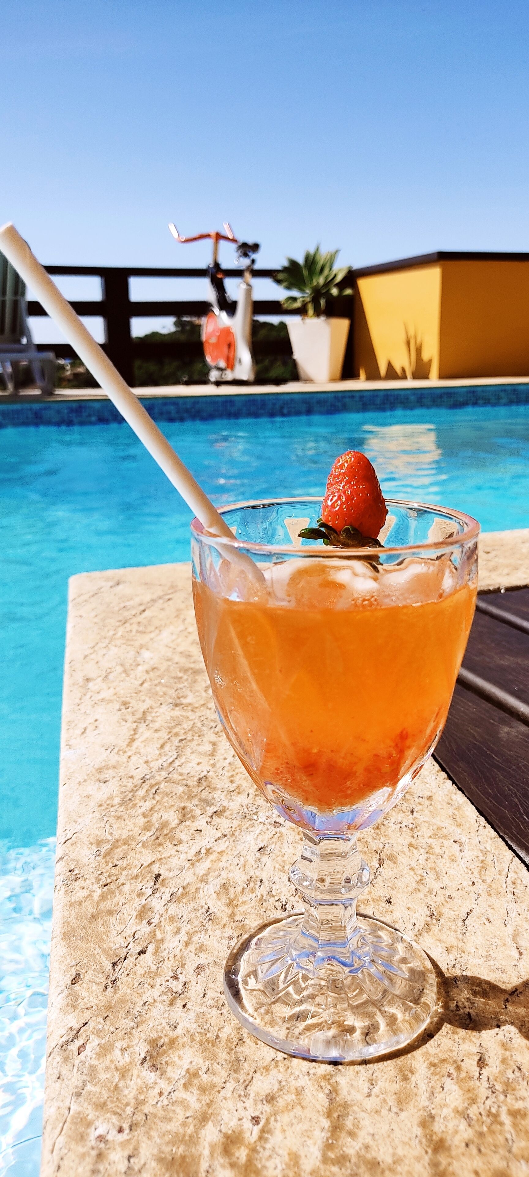 Una copa de cóctel con una fresa en el borde, sobre una mesa junto a una piscina.
