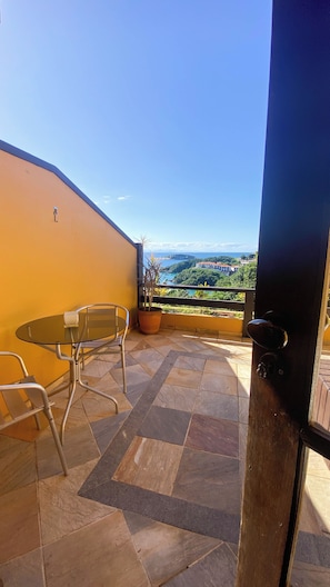 Ein Balkon mit Tisch und Stühlen, einem Blumentopf und Blick auf eine Küstenlandschaft.