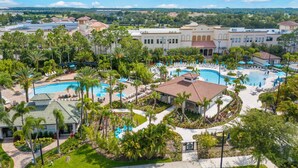 A resort with multiple swimming pools, palm trees, and a large building with a red-tiled roof. - VCR4-308 - 3BR Condo In Vista Cay near Disney (Orlando)