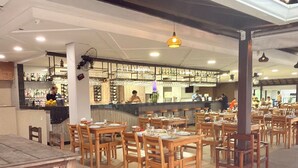 Een ruim restaurant met meerdere houten tafels en stoelen, een bar met flessenkasten, en een hoge plafond met hangende lampen.