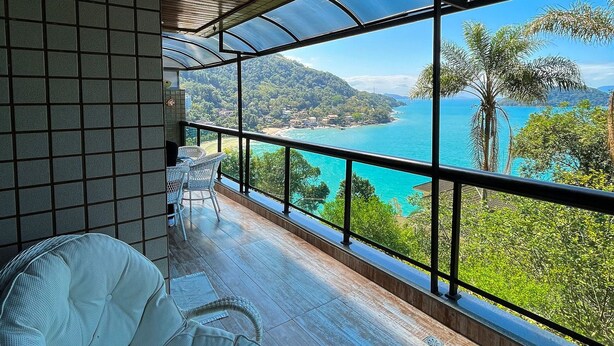 Een balkon met uitzicht op een turquoise zee en groene heuvels, houten vloer en een witte rieten stoel.