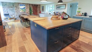 A modern kitchen with a large island, wooden flooring, and a stone accent wall. - Flaska House (Penrith)