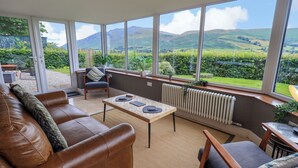 A sunlit room with a large window, a brown leather sofa, a wooden table, and a chair. - Flaska House (Penrith)