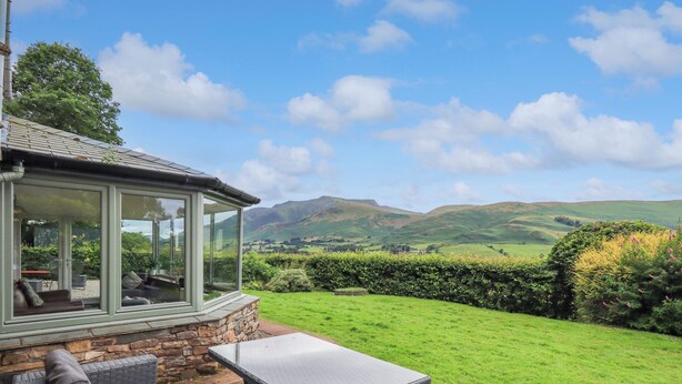 A patio with a table and chairs, a glass-enclosed seating area, and a view of rolling hills. - Flaska House (Penrith)