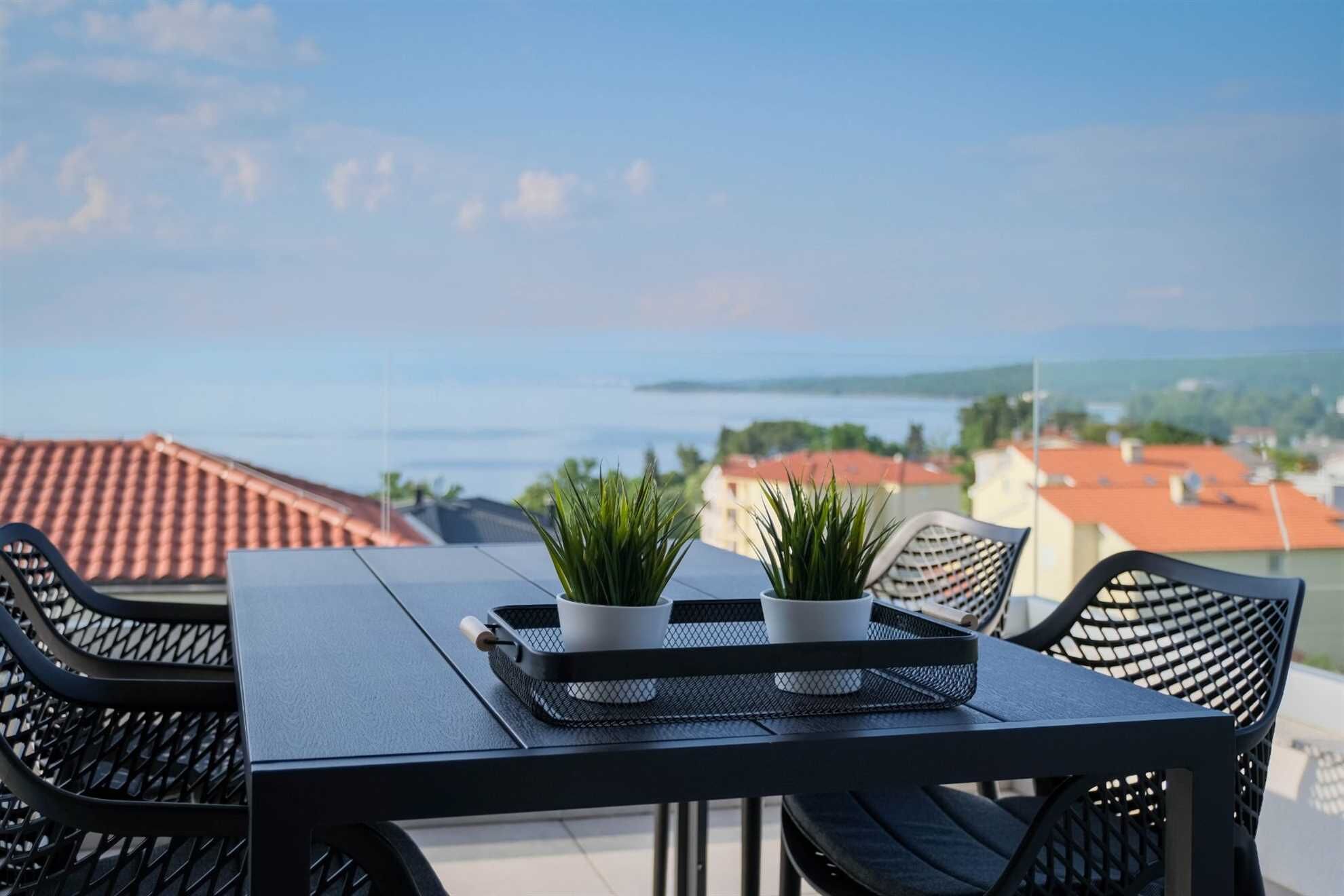 A terrace with a table and chairs, two potted plants, and a view of the sea and buildings in the background.