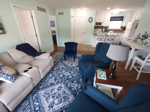 Uma sala de estar com sofá, uma poltrona azul, tapete estampado e um espaço de jantar com mesa e cadeiras.