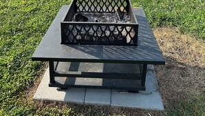 A square fire pit with a lattice design on a stone platform, surrounded by a grassy field and trees, with a mountain in the background.