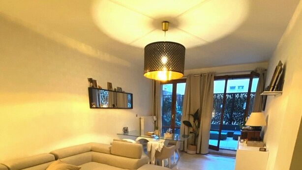 A modern living room with a sectional sofa, a glass coffee table, and a pendant light. There is a dining area with a small table and chairs. The room has a large window with a view of the outdoors. - Appart Avec Garage et Terrasse Thier Buffon (Avignon)